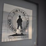 A metallic sign on a door has a silhouette of a soldier holding a gun. A circle goes around the soldier with the words "Virtual Soldier Research."