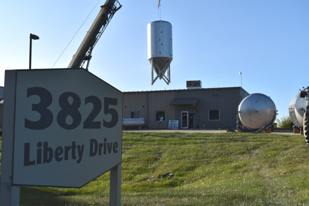 In the foreground is a sign that says "3825 Liberty Drive." In the background is a warehouse, and a large beer fermenter is being lifted in the air by a crane.