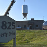 In the foreground is a sign that says "3825 Liberty Drive." In the background is a warehouse, and a large beer fermenter is being lifted in the air by a crane.