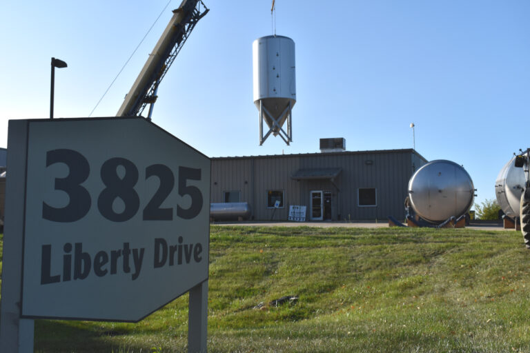In the foreground is a sign that says "3825 Liberty Drive." In the background is a warehouse, and a large beer fermenter is being lifted in the air by a crane.