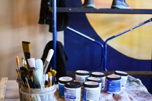 Painting supplies can be seen on a table, as Candice Warby paints an indoor mural. 