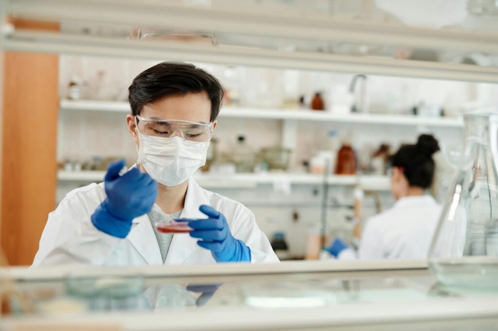 A scientist wearing a face mask and lab coat holds a petri dish.