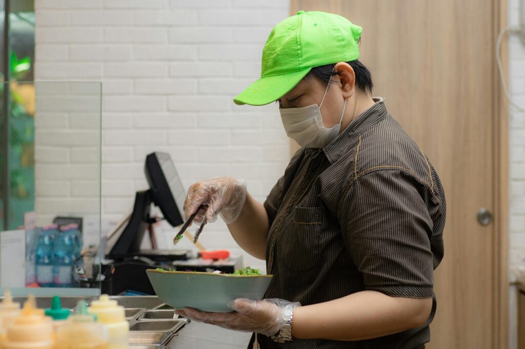 A masked restaurant worker stands behind a counter holding a bowl of food.