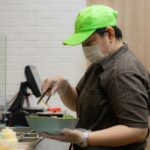 A masked restaurant worker stands behind a counter holding a bowl of food.