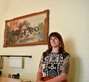 State of Mind owner Carissa Starleaf poses with the store sign, a repurposed thrifted painting emblazoned with "State of Mind" across the middle. CREDIT ANNIE SMITH BARKALOW