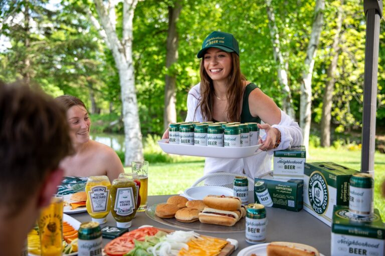 A smiling woman brings a tray of Neighborhood Beer to some women sitting at a picnic table.