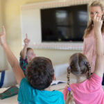 Rear view of a diverse group of schoolchildren sitting at tables in an elementary school classroom and raising their hands to answer their Caucasian female teacher, standing by the whiteboard in the background